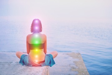 Portrait of young beautiful woman sits in a pose of half lotus practicing yoga meditation at blue summer sea environment glowing all seven chakra Kundalini energy exercise Back view Inner mind consept - obrazy, fototapety, plakaty
