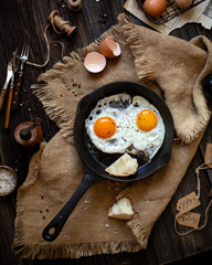 Overhead shot of cast-iron pan with two fried eggs and bread on sackcloth standing on wooden rustic...