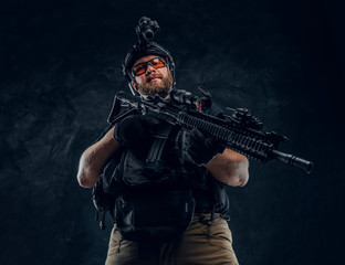 Special forces soldier wearing body armor and helmet with night vision holding an assault rifle. Studio photo against a dark textured wall