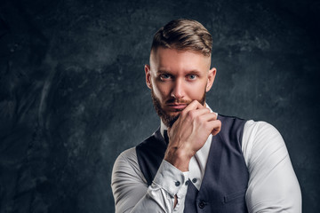 Closeup portrait of an elegantly dressed young man with stylish beard and hair holding hand on a chin and looking at a camera. Studio photo against a dark wall background