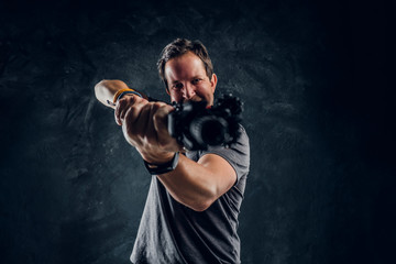 Fototapeta na wymiar Distraught middle-aged man dressed in casual clothes holding an assault rifle and aims at the target. Studio photo against a dark textured wall
