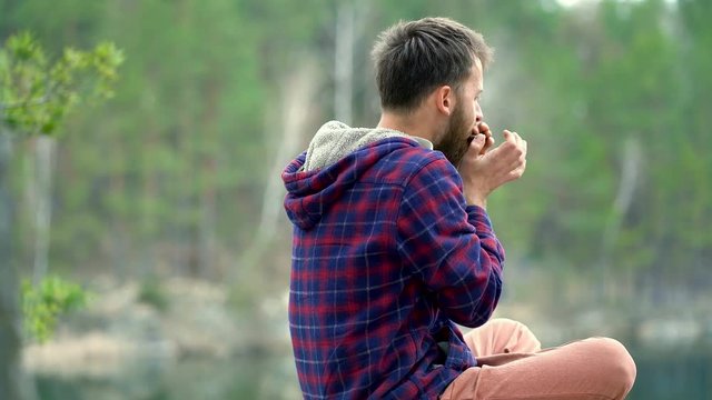 Young beard man playing on the harmonica. Sitting on the rock near the lake. Steadycam shot.