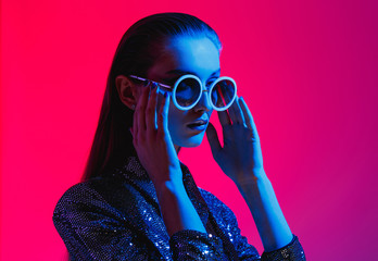 Fashion girl with long hair and round sunglasses in a black shining dress poses in neon light in...