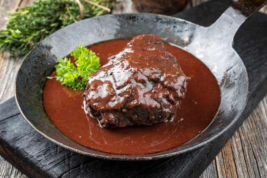 Traditionelle geschmorte Ochsenbacke in brauner Rotwein Sauce mit Kräutern als closeup in einer gusseisernen Pfanne