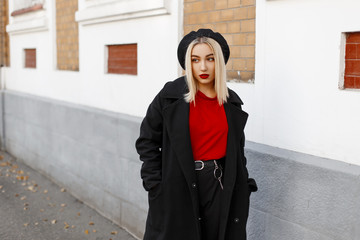 Fashionable pretty modern young blond woman in a stylish retro-style outerwear with red lips in a beret walks down the street on an autumn afternoon near the building. Cute girl enjoying the weekend.