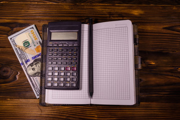 Opened notepad, one hundred dollar banknotes, scientific calculator and pencil on wooden table. Top view