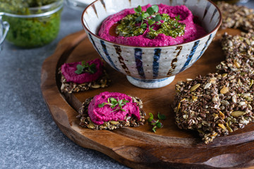 Roasted beet hummus in ceramic bowl, flax seed crackers on wooden cutting board, pesto sauce jar -...