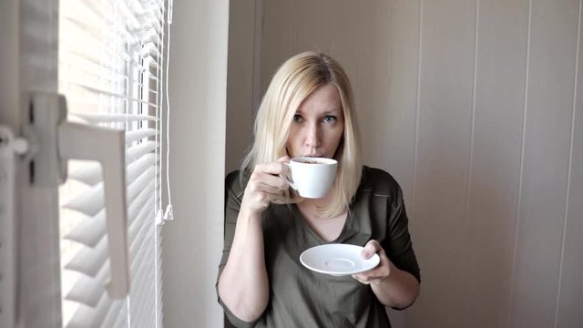 Young sad thinkful beautiful blond woman standing near the window with blinds in the morning and drinking coffee