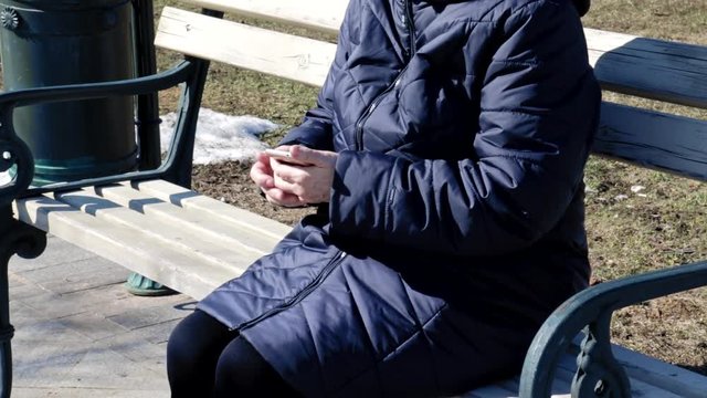 A senior elderly woman sitting on a bench in park outdoors and using tapping smartphone -she is useing smartphone to connect with people on social, texting messages ang searching internet
