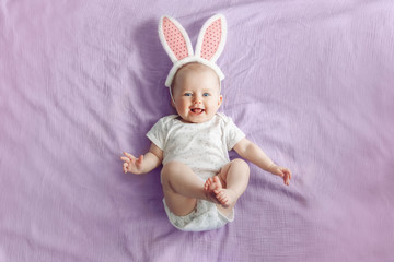 Cute adorable white Caucasian baby girl wearing pink Easter bunny ears lying on pink purple bed in...