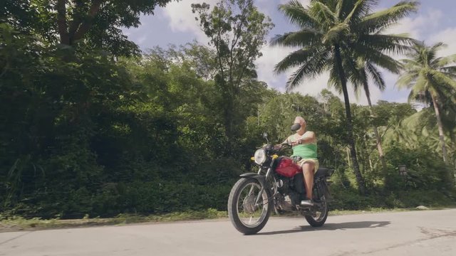 Man motorcyclist driving on motorbike on road with green palm trees landscape. Mature man riding motorcycle on road in tropical palm while summer travel. Moto trip.