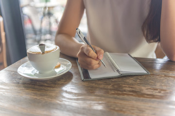 Female hand writing note beside cup of hot cappuccino
