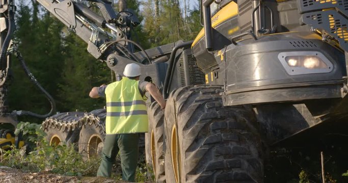 The operator of the logging machine to cutting trees in white helmet and green jumpsuit went into the forest to inspect equipment after work