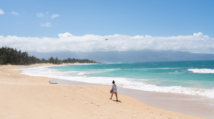 Asian woman walking across the tropical sandy beach with turquoise waves crashing on it. Plane flies away in the background.