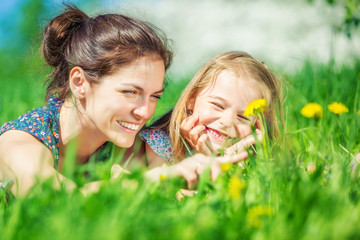 Young woman and her daughter lying on green summer grass with blooming yellow dandellions