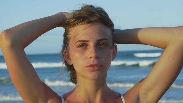 4K close-up portrait of beautiful young woman with hands on head looking into camera on the beach with waves in the background, South Africa