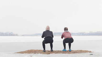 Two slim women standing on the snowy beach and doing squats