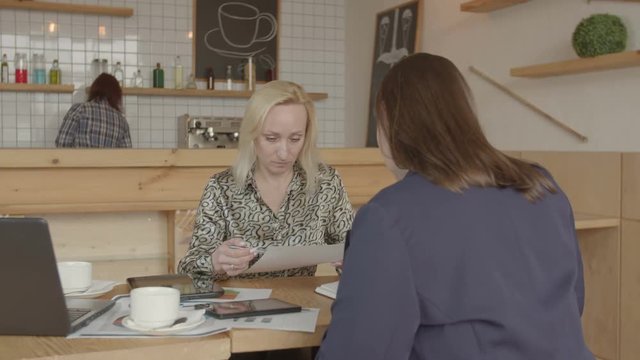 Successful female small business owner meeting with insurance agent in cafeteria intending to insure property. Woman professional broker offering entrepreneur insurance policy template for review.