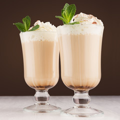 Coffee in two goblets with whipped cream, chocolate powder, fresh green mint in brown modern restaurant interior, closeup, square.