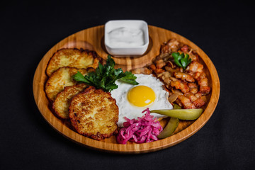 Potato pancakes with a fried egg, bacon, sourcream, horse radish, pickles and parsley on a wooden plate on black background