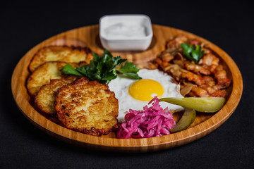 Potato pancakes with a fried egg, bacon, sourcream, horse radish, pickles and parsley on a wooden plate on black background