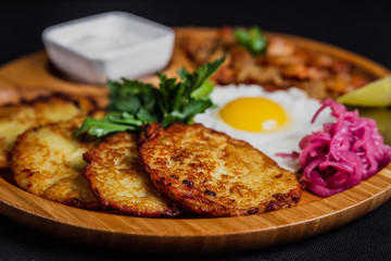 Potato pancakes with a fried egg, bacon, sourcream, horse radish, pickles and parsley on a wooden plate on black background
