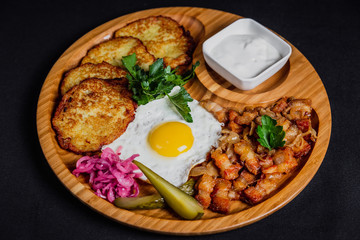 Potato pancakes with a fried egg, bacon, sourcream, horse radish, pickles and parsley on a wooden plate on black background