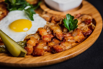 Potato pancakes with a fried egg, bacon, sourcream, horse radish, pickles and parsley on a wooden plate on black background