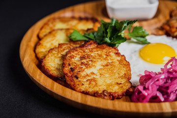 Potato pancakes with a fried egg, bacon, sourcream, horse radish, pickles and parsley on a wooden plate on black background