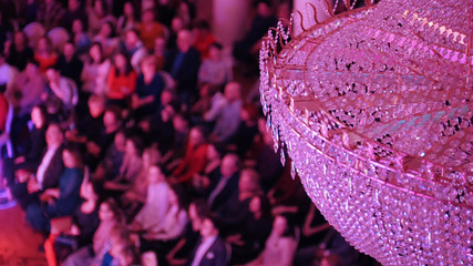A concert in the concert hall with a big crystal chandelier. People sitting on the chairs