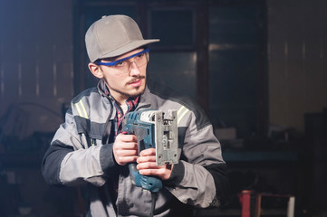 Portrait of a young carpenter joiner in overalls wearing a cap and goggles with an electric jigsaw...