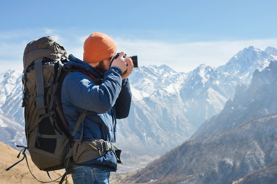 Portrait of a bearded male photographer in sunglasses and a warm jacket with a backpack on his back and a reflex camera in his hands takes pictures against the background of snow-capped mountains