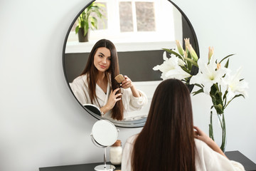 Beautiful young woman combing her healthy long hair near mirror at home