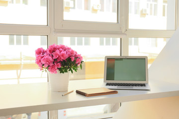 Pot with beautiful blooming azalea, laptop and notebook on table near window