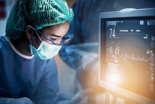 Team of doctors or surgeons with electrocardiogram monitor in hospital surgery operating emergency room showing patient heart rate, medical concept