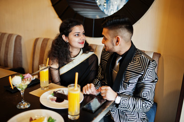 Lovely indian couple in love, wear at saree and elegant suit, sitting on restaurant. On table sweet desserts and juices.