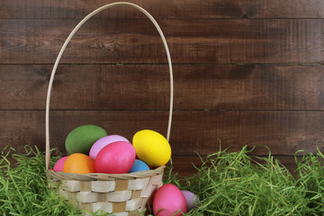 Colorful eggs in basket on green grass against wooden wall. Easter background or egg hunt concept. Pink, yellow, green, orange eggs, hay