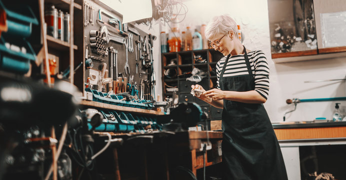 Beautiful Caucasian female worker with short blonde hair and eyeglasses taking tool fromm wall to repair bicycle. Bike workshop interior.