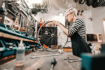 Cute Caucasian female worker holding and repairing bicycle wheel while standing in bicycle workshop.