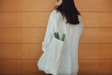 Back view of a young girl with a tote cotton blank reusable bag for shopping with leek and fresh vegetables inside, wooden wall on the background, concept of healthy farm sustainable shopping