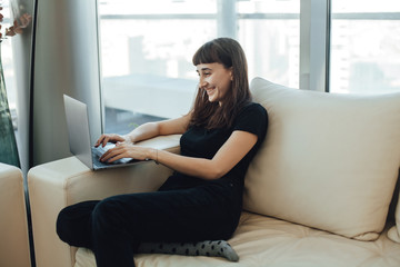Obraz premium Happy smiling hipster girl typing on a laptop computer keyboard, chatting with family or friends via online messenger while sitting on a cozy sofa ot home with modern interior design
