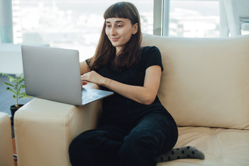 Naklejka premium Happy pretty casual dressed woman sitting on a sofa at home and browsing internet via laptop computer for distant work or study or doing online shopping and watching videos
