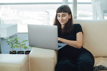 Happy young girl watching online movies and videos or doing online shopping using her modern laptop with wifi while sitting on a sofa at home, woman browsing internet via computer laptop at her house