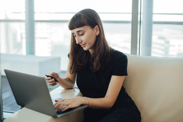 Happy smiling hipster girl using modern laptop computer and credit card for online shopping while sitting on a sofa at home, concept of doing shopping online with the delivery right to your door