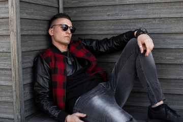 Young hipster man in a trendy checkered red jacket with leather sleeves in dark sunglasses in a...