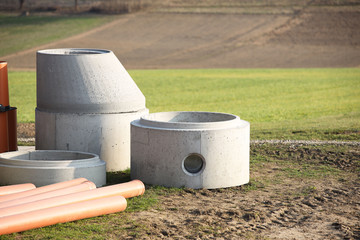 Elements of concrete structures for the arrangement of sewage systems lie on the green grass in the rays of the sun. The walls of the mine made earth wells for the wiring of various networks under the