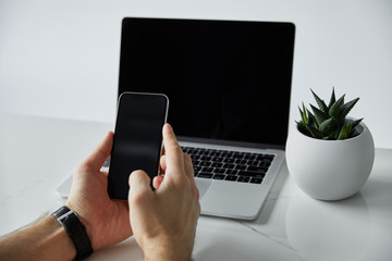 partial view of man using smartphone near laptop with blank screen, and flowerpot pot isolated on grey