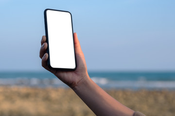 Mockup image of woman's hand holding black mobile phone with blank desktop screen by the beach and sea with blue sky background