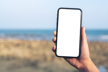 Mockup image of woman's hand holding black mobile phone with blank desktop screen by the beach and...