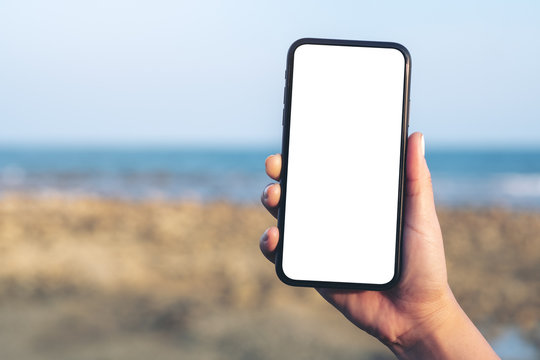 Mockup image of woman's hand holding black mobile phone with blank desktop screen by the beach and sea with blue sky background